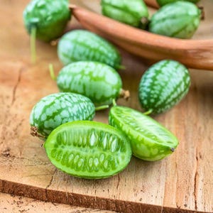 May include: A collection of small, green, oval-shaped cucamelons, some whole and others sliced open to reveal the juicy interior. The cucamelons have a light green, speckled exterior and are displayed on a wooden surface.