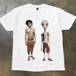 Puede incluir: Una camiseta blanca de manga corta con un estampado gráfico de una persona con cabello oscuro y rizado, una toalla blanca alrededor del cuello y pantalones cortos naranjas, junto a una figura alienígena con chaleco y pantalones cortos naranjas.