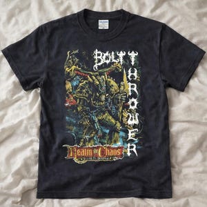 May include: A black short-sleeve t-shirt featuring a graphic print of a horned, armored figure holding a weapon. The text "BOLT THROWER" is printed vertically in white, and "REALM OF CHAOS" appears in a stylized banner at the bottom of the design.