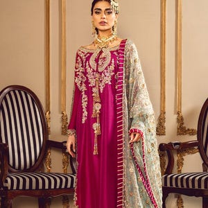 May include: A magenta-colored formal dress with gold embroidery and a matching sheer scarf. The dress features long sleeves and intricate detailing. The scarf is light blue with gold accents and a pink border. The background includes two chairs.