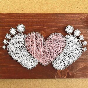 Puede incluir: Una placa de madera con acabado marrón oscuro presenta un diseño de arte de hilo. El diseño incluye dos huellas de bebé que flanquean un corazón rosa. Las huellas y el corazón están hechos de hilo blanco y rosa.