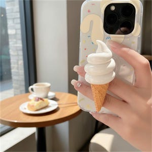 May include: Clear phone case with a white ice cream cone grip. The case has a light beige bear design and colorful confetti. The ice cream cone is white with a waffle cone base. The phone is held in a person's hand.