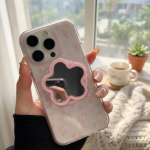 Könnte beinhalten: Transparente Handyhülle mit rosa Blumenmuster und einem rosa blütenförmigen Spiegel. Die Hülle hat einen Kameraausschnitt. Der Hintergrund umfasst ein Fenster, eine kleine Pflanze und eine Tasse Tee.