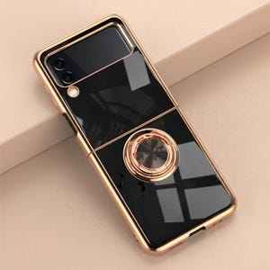 May include: A black and gold phone case with a ring holder. The case has a glossy black finish with gold-coloured trim and a circular ring holder in the centre. The phone case is designed to protect the phone and provide a secure grip.