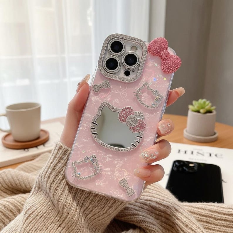 K&ouml;nnte beinhalten: Rosa Handyh&uuml;lle mit Hello Kitty-Design. Die H&uuml;lle hat einen Spiegel, Strassdetails und eine Schleife. Die Kameralinse ist von Strass umgeben. Die H&uuml;lle hat ein gl&auml;nzendes Finish.