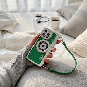 May include: A green and silver phone case designed to resemble a vintage camera. The case has a green leather-like texture on the back and a green strap. The case has the text "camera", "35-70mm", "1-photo passion", and "4880W".