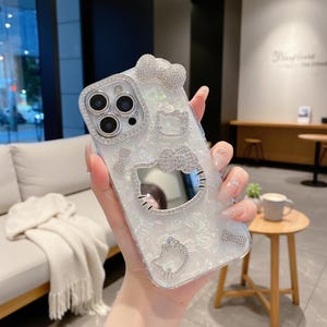 K&ouml;nnte beinhalten: Eine wei&szlig;-silberne Handyh&uuml;lle mit Perlmutt-Finish, verziert mit glitzernden Strasssteinen. Die H&uuml;lle hat eine Schleife, Hello Kitty-Designs und einen Kamerarahmen. Das Design ist mit mehreren Strasssteinen verziert.