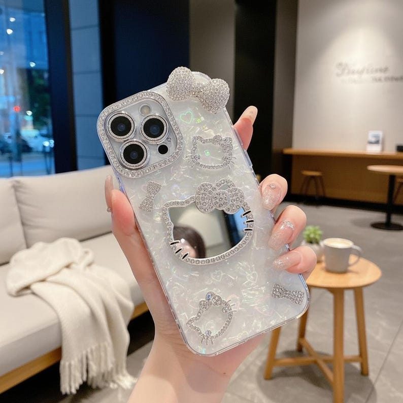 K&ouml;nnte beinhalten: Eine wei&szlig;-silberne Handyh&uuml;lle mit Perlmutt-Finish, verziert mit glitzernden Strasssteinen. Die H&uuml;lle hat eine Schleife, Hello Kitty-Designs und einen Kamerarahmen. Das Design ist mit mehreren Strasssteinen verziert.