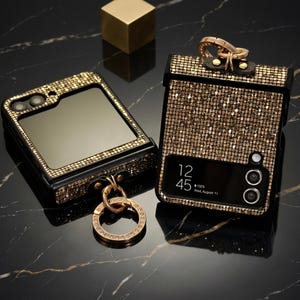 Può includere: Due custodie per telefono impreziosite con dettagli oro e neri. Una custodia è aperta, rivelando uno schermo, mentre l'altra è chiusa, mostrando uno schermo con l'ora 12:45 e la data mercoledì 11 agosto. Entrambe le custodie hanno un anello dorato.