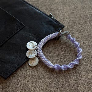 May include: A lavender and white braided bracelet with three small, round charms. The charms have the letters "O", "R", and "E" engraved on them. A black leather pouch is partially visible in the background.