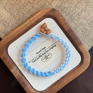 May include: A blue and white braided bracelet with a brown butterfly clasp. The bracelet is displayed on a white card inside a wooden tray. The card has text that reads "French Paris I met you The dead tree of spring."