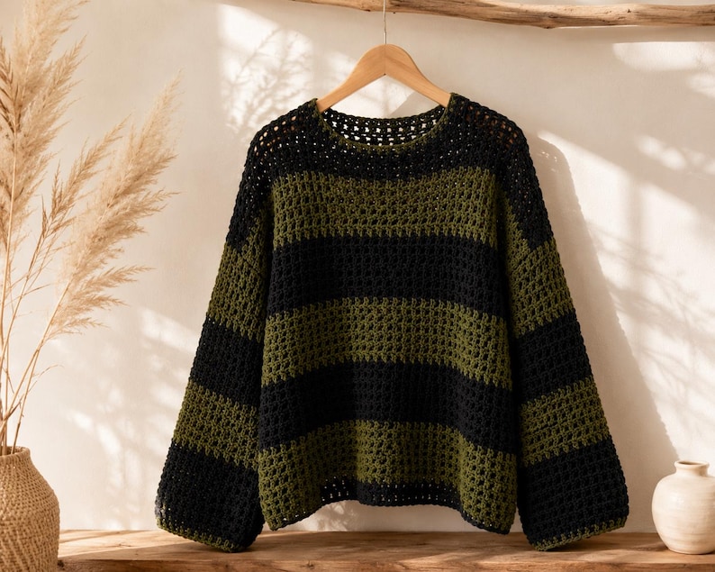 Puede incluir: Un su&eacute;ter de manga larga con un patr&oacute;n de rayas horizontales en colores negro y verde oliva alternados. La prenda est&aacute; confeccionada con un punto de ganchillo abierto, cuello redondo y un corte holgado, colgado de una percha de madera.