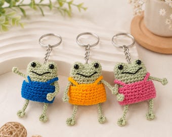 Puede incluir: Tres llaveros de rana hechos a ganchillo, cada uno con un cuerpo verde claro, ojos de cuentas negras y una boca bordada. Las ranas llevan pequeños vestidos de colores sólidos en azul, amarillo y rosa, y están unidas a anillas de metal plateado.