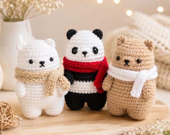 Puede incluir: Tres pequeños osos de ganchillo colocados uno al lado del otro. El oso de la izquierda es blanco con bufanda beige, el central es un panda blanco y negro con bufanda roja, y el de la derecha es beige con bufanda blanca. Tienen ojos y nariz negros.