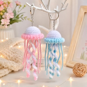 May include: Two handmade crochet jellyfish keychains. One has a pink and white body with pink tentacles, the other has a blue and white body with blue tentacles. Both have a white top and silver key rings.