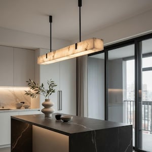 May include: A rectangular pendant light fixture with a translucent, marbled stone-like shade suspended by two thin black rods from the ceiling. The light emits a warm glow, illuminating the kitchen island below with its linear, elongated design.