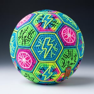 May include: A colorful, crocheted ball with a geometric pattern. The ball features hexagonal panels in shades of blue, green, pink, and yellow. Each panel has embroidered designs, including lightning bolts and circuit board patterns. A small tag reads "NEON IMPACT".