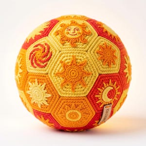May include: A crocheted ball with a soccer ball design. The ball features a variety of sun and moon designs in shades of yellow, orange, and red. The ball has a tag that reads "The Brightest Kick".