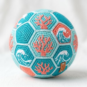 May include: A crocheted soccer ball featuring a sea-themed design. The ball is primarily turquoise with coral and white accents. Hexagonal panels display coral, waves, and the text "Summer Splash" on a small leather tag.