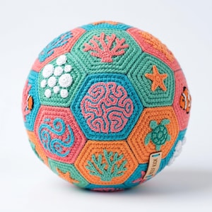 May include: A colorful, crocheted ball featuring a variety of sea life designs. The ball is composed of hexagonal panels in shades of teal, coral, orange, and white. Designs include coral, starfish, and a turtle. A small tag reads "Reef Kick".