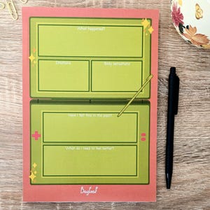 May include: A pink and green journal with prompts like "What happened?" and "Emotions". The design resembles a retro video game. A black pen and a floral mug are on a wooden surface.