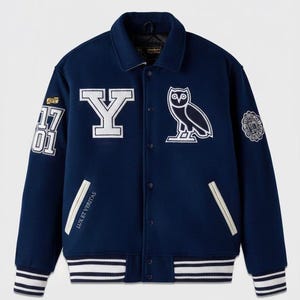 May include: Navy blue varsity jacket with white accents. Features a large white "Y" and an owl patch. Includes patches with the numbers "17 01" and the text "LUX ET VERITAS". The jacket has a striped collar, cuffs, and hem.