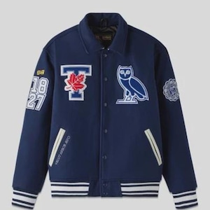 May include: A navy blue varsity jacket with white and red embroidered patches. The jacket features an owl, a maple leaf, and the numbers "18 27". White stripes adorn the cuffs and hem. The text "VELUT ARBOR AVO" is visible.