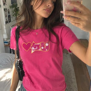 Op de afbeelding: Een felroze T-shirt met korte mouwen en een grafisch ontwerp op de borst. Het ontwerp bevat een effen roze hart, een geel omrande ster met een realistisch menselijk oog en de letter U met kleine witte steraccenten.