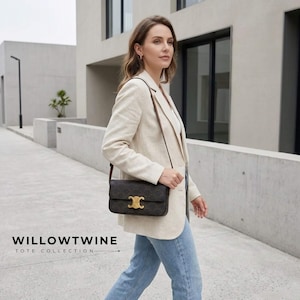 May include: A dark brown rectangular shoulder bag with a repeating monogram pattern and a gold-toned metal clasp on the front flap. The bag features a thin, adjustable shoulder strap and is worn over a light beige blazer and blue denim jeans.