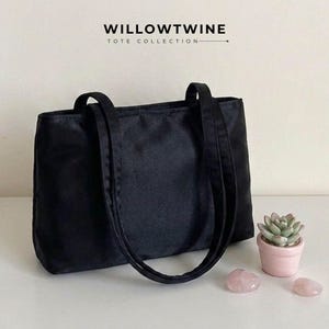 May include: A black tote bag with two shoulder straps, featuring the text WILLOWTWINE TOTE COLLECTION at the top. The bag is positioned next to a small pink pot containing a green succulent plant and two smooth, light pink stones.