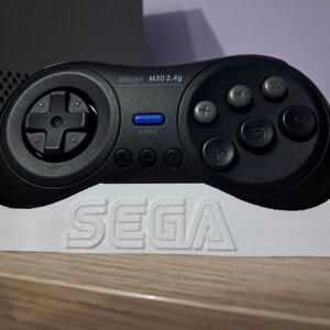 Könnte beinhalten: Schwarzer kabelloser Controller mit grauen Tasten, einer blauen Starttaste und einem Steuerkreuz. Der Controller ruht auf einem weißen Ständer mit dem SEGA-Logo. Es ist ein M30 2.4g Modell.
