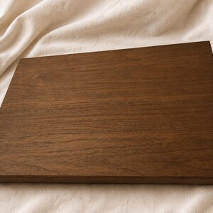 May include: A rectangular wooden chopping board in a deep brown hue. The wood grain is clearly visible, providing texture. The board is placed on a neutral, off-white fabric background.