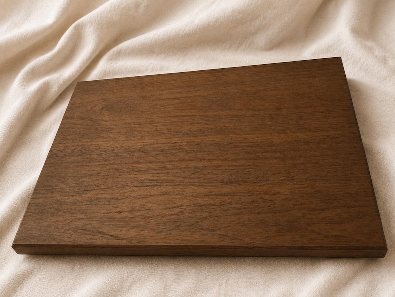May include: A rectangular wooden chopping board in a deep brown hue. The wood grain is clearly visible, providing texture. The board is placed on a neutral, off-white fabric background.