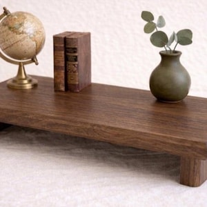 Puede incluir: Un soporte de monitor de madera marrón oscuro con un pequeño globo de latón, dos libros antiguos y un pequeño jarrón verde con vegetación. El soporte es rectangular con un borde elevado y se apoya en cuatro patas cortas.