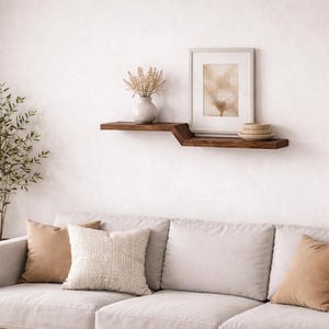 May include: A living room scene featuring a light-colored sofa with decorative pillows. A dark wood shelf displays a framed print, a vase with dried flowers, and small bowls. A floor lamp and a potted plant add to the decor.