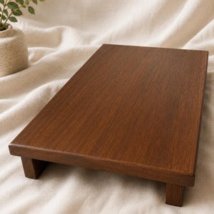May include: A rectangular, dark brown wooden serving tray with a raised edge and short legs. The tray is made of a rich, warm-toned wood and sits on a cream-colored fabric surface. The tray is ideal for serving food or displaying decorative items.