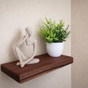 May include: A dark wood floating shelf with a light-colored statue of a person in a seated pose. A small white pot with green plants sits on the shelf. The shelf is mounted on a textured beige wall.