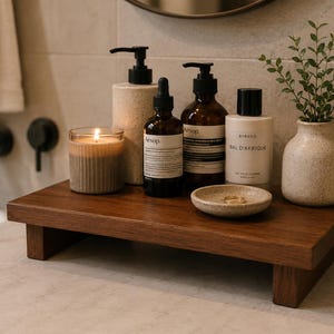 May include: A dark wooden bathroom shelf displays a lit candle, several brown bottles with black pumps and labels, a small ceramic dish with rings, and a small vase with greenery. The shelf is a dark brown colour.
