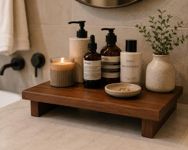 May include: A dark wooden bathroom shelf displays a lit candle, several brown bottles with black pumps and labels, a small ceramic dish with rings, and a small vase with greenery. The shelf is a dark brown colour.
