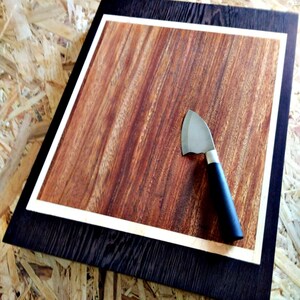 Peut inclure: Une planche à découper en bois avec un cadre marron foncé et une bordure en bois clair. La surface de coupe est d'un brun rougeâtre riche avec un motif de grain de bois naturel. Un petit couteau à lame incurvée avec un manche noir repose sur la planche.
