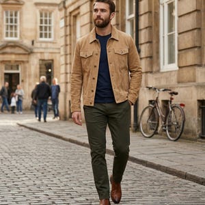 May include: A man wearing a tan suede jacket, navy blue t-shirt, and olive green pants walks on a cobblestone street. He is also wearing brown leather boots. A bicycle is parked in the background.