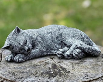 May include: A gray cat statue in a sleeping position, with its eyes closed and paws tucked in. The cat is resting on a tree stump, with a blurred green background. The statue has a textured surface and detailed features.