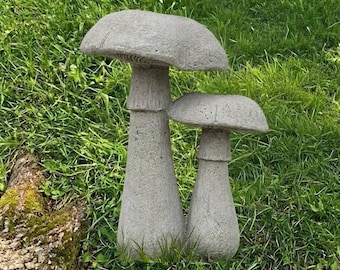 May include: Two gray concrete mushroom sculptures of varying heights, each featuring a wide, rounded cap with visible gills underneath and a thick, cylindrical stem. The textured, stone-like surface of the garden ornaments is uniform in color.