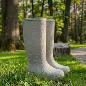 May include: A pair of tall, light gray rubber boots with a textured, matte finish. The boots feature a rounded toe, a defined heel, and subtle molded lines around the ankle and calf area, standing upright in a grassy outdoor environment.
