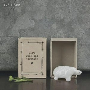 May include: A small, open matchbox containing a white ceramic bear figurine. The matchbox is tan and features the text "Let's grow old together" and a heart symbol. The box measures 4.5 x 3 cm.