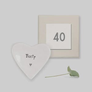 Könnte beinhalten: Eine weiße herzförmige Keramikschale mit dem Aufdruck "Forty" und einem kleinen Herzsymbol. Neben der Schale befindet sich eine quadratische Tafel mit der Zahl "40". Ein kleines grünes Blatt ist ebenfalls vorhanden.
