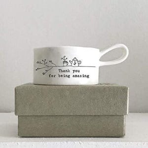 Peut inclure: Une tasse en céramique blanche avec une anse incurvée, ornée d'un dessin au trait noir d'une plante, d'une théière et du texte "Thank you for being amazing". La tasse est posée sur une boîte rectangulaire grise.