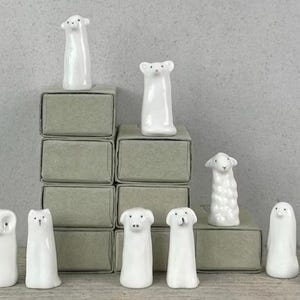 Puede incluir: Una colección de pequeñas figuras de animales de cerámica blanca, que incluyen un oso, una oveja, un búho, un gato, un perro y un fantasma. Las figuras se exhiben en pequeñas cajas grises apiladas. El fondo es una pared gris claro.