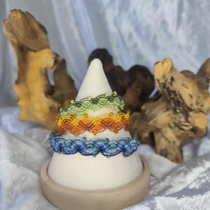May include: A display of handmade macrame bracelets in shades of blue, orange, green, and yellow. The bracelets are stacked on a white cone-shaped display stand. The background features a textured, light-colored fabric and decorative wooden elements.