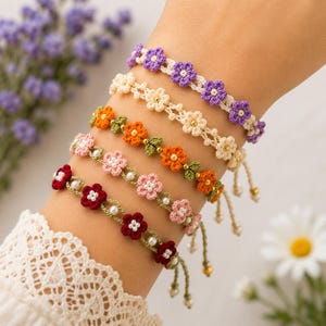 May include: A set of five handmade crochet bracelets featuring floral patterns in purple, cream, orange, light pink, and dark red. Each bracelet is accented with small beads and includes adjustable tie closures with dangling bead ends.